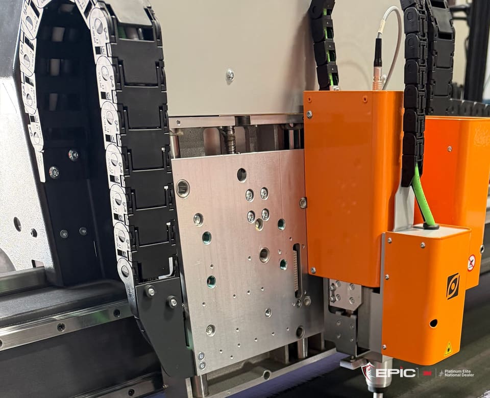 The Epic Precision cutting head poised over a sheet of clear 3M Prestige 40 on the cutting table in Epic's Aurora facility, the warehouse reflected in the film