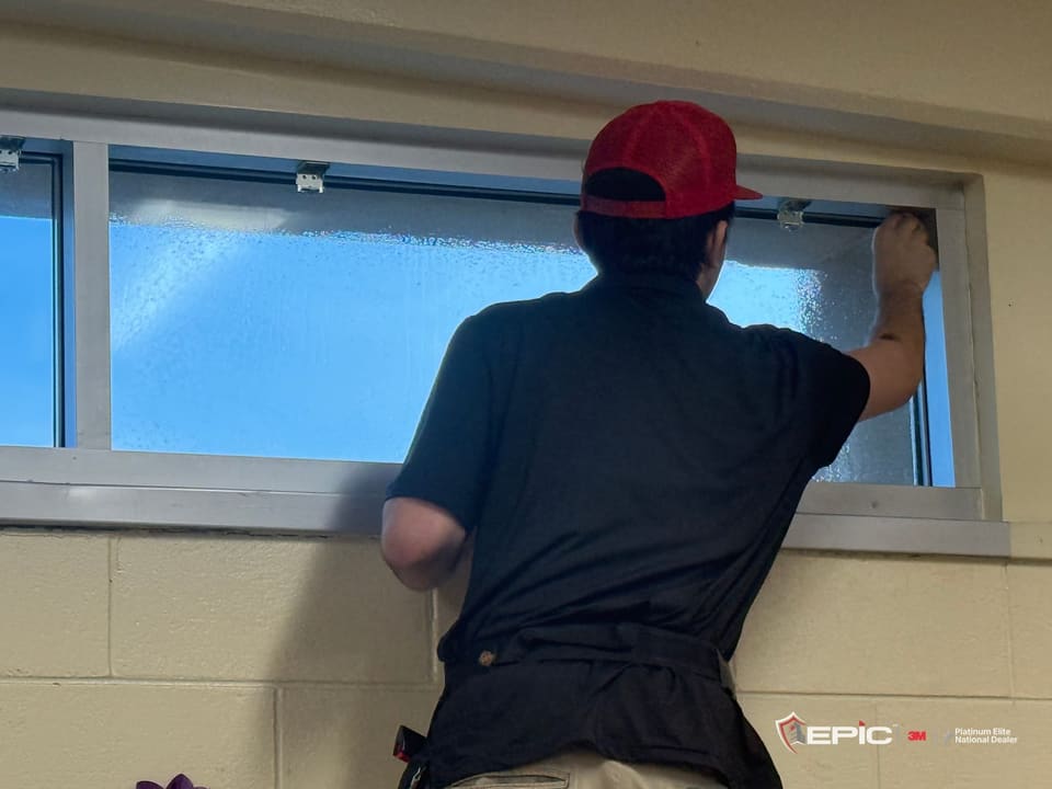 A Certified Installation Specialist in a red Epic cap working 3M S2400 onto a band of clerestory glass, back to the camera