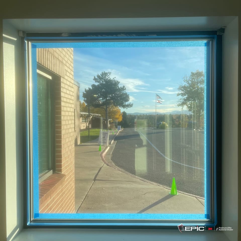 A square entry sidelite from inside, the film on and every edge of the frame taped in blue, a flagpole and the mountains through the glass