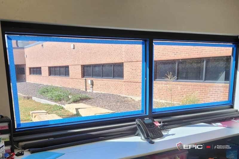 Two school windows with blue painter's tape run around the inside of each frame, brick building and gravel bed outside