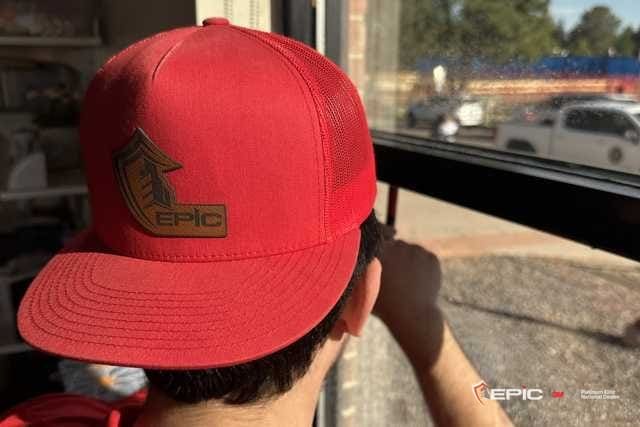Certified Installation Specialist in a red Epic cap scraping a classroom window from inside, trees and a parking lot beyond the glass