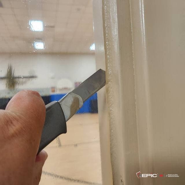 A blade run along the edge of a white window frame next to the glass, a school gym floor and backboard visible through the window