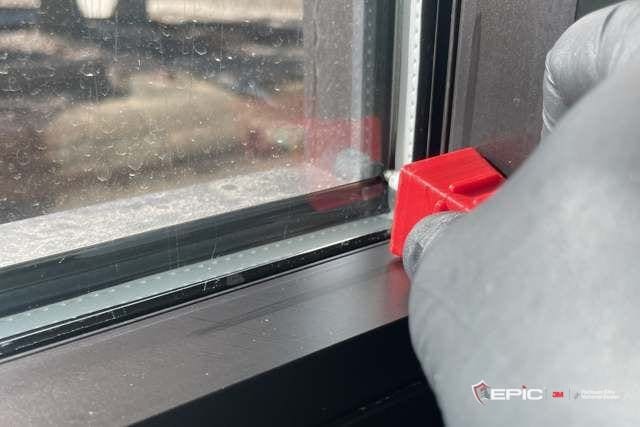 Gloved hand holding a red block tool at the corner where the glass meets the dark window frame