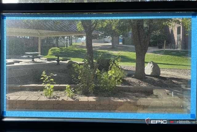Lower lite of a classroom window with blue tape run around the perimeter marking the line for the 3M IPA bead; courtyard, picnic tables and a shade structure outside