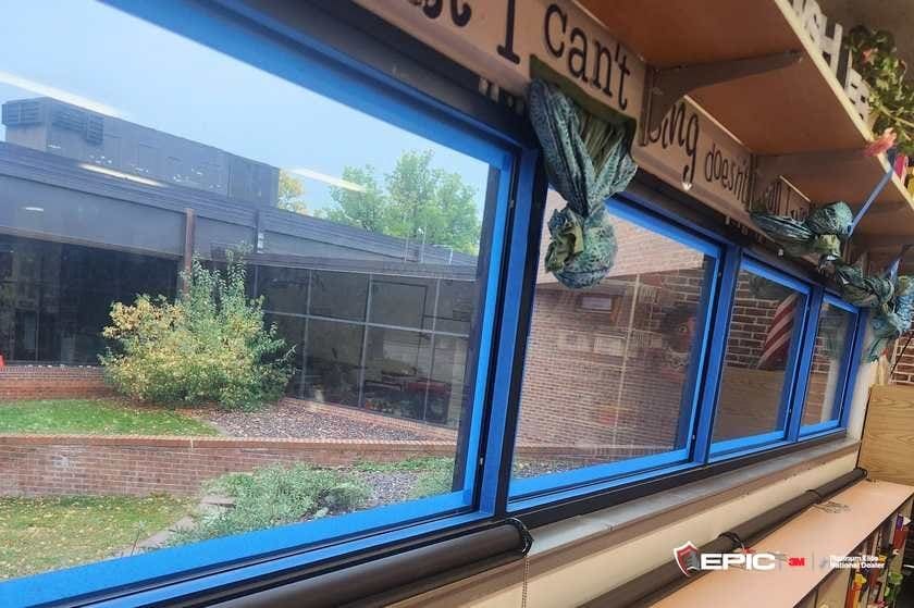 A row of library windows taped blue at the frames, roller shades on the sill, courtyard and brick wing outside