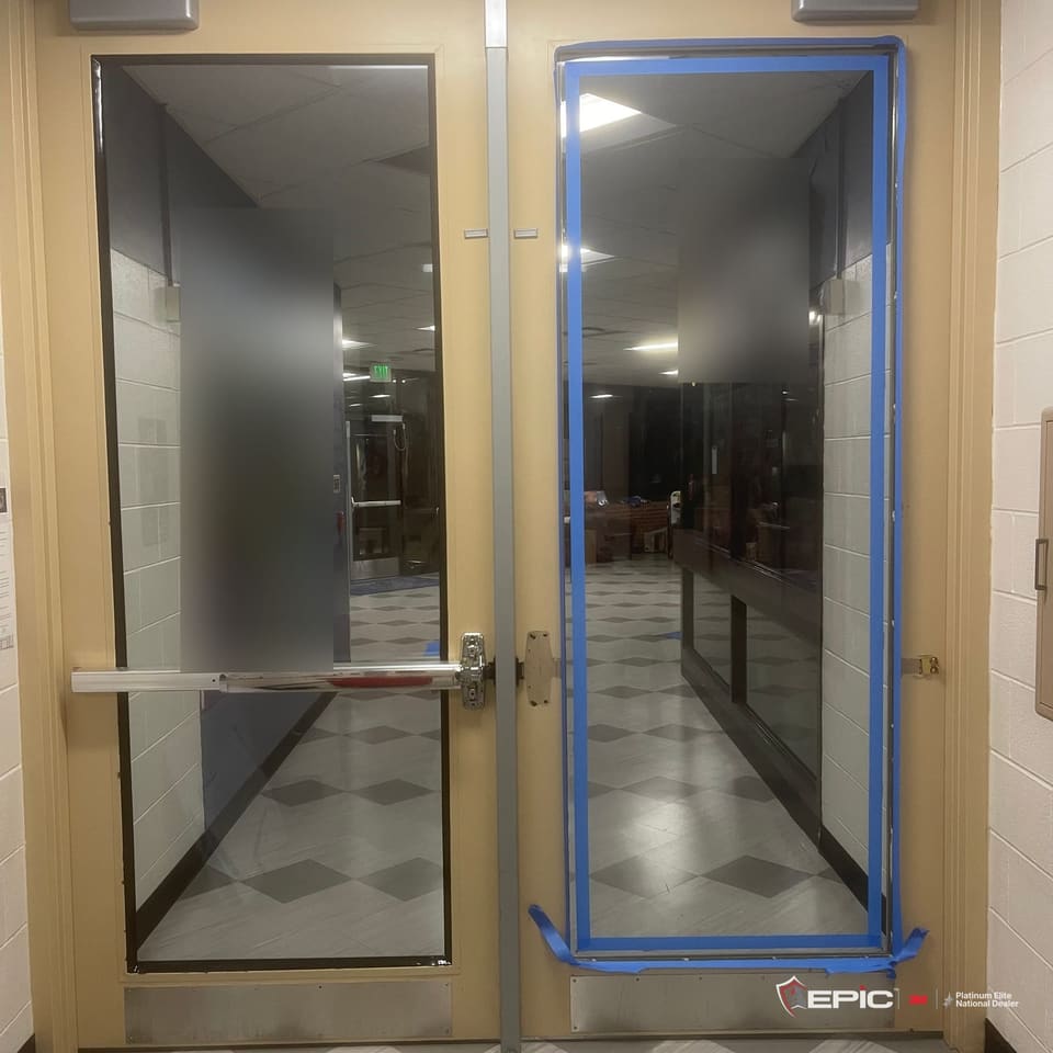 A pair of school entry doors from the hallway: the left lite filmed with the 3M IPA bead at the frame, the right lite filmed and taped in blue on every edge