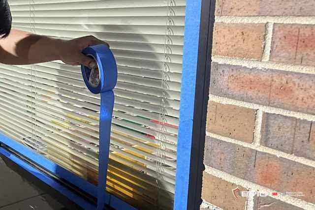 Blue tape run along a window frame over the blinds, a roll of tape in hand