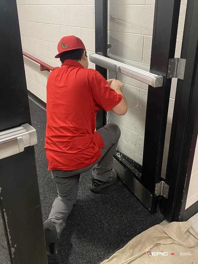 A Certified Installation Specialist in a red Epic shirt kneeling at a school vestibule door, working film onto the glass below the push bar