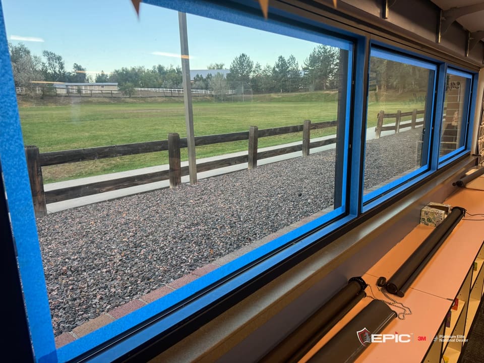 Three school windows over a low counter, blue tape run around every frame, a split-rail fence and field outside