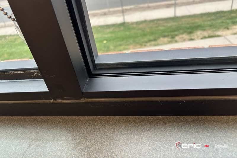 The sill corner of a dark classroom window frame with the glazing gasket removed and the glass filmed, grass and a fence outside