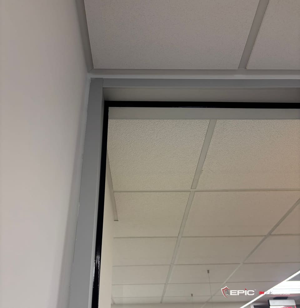 The corner of a full-height glass sidelite beside a maple office door, tape off, the black 3M Impact Protection Attachment bead running down the frame edge