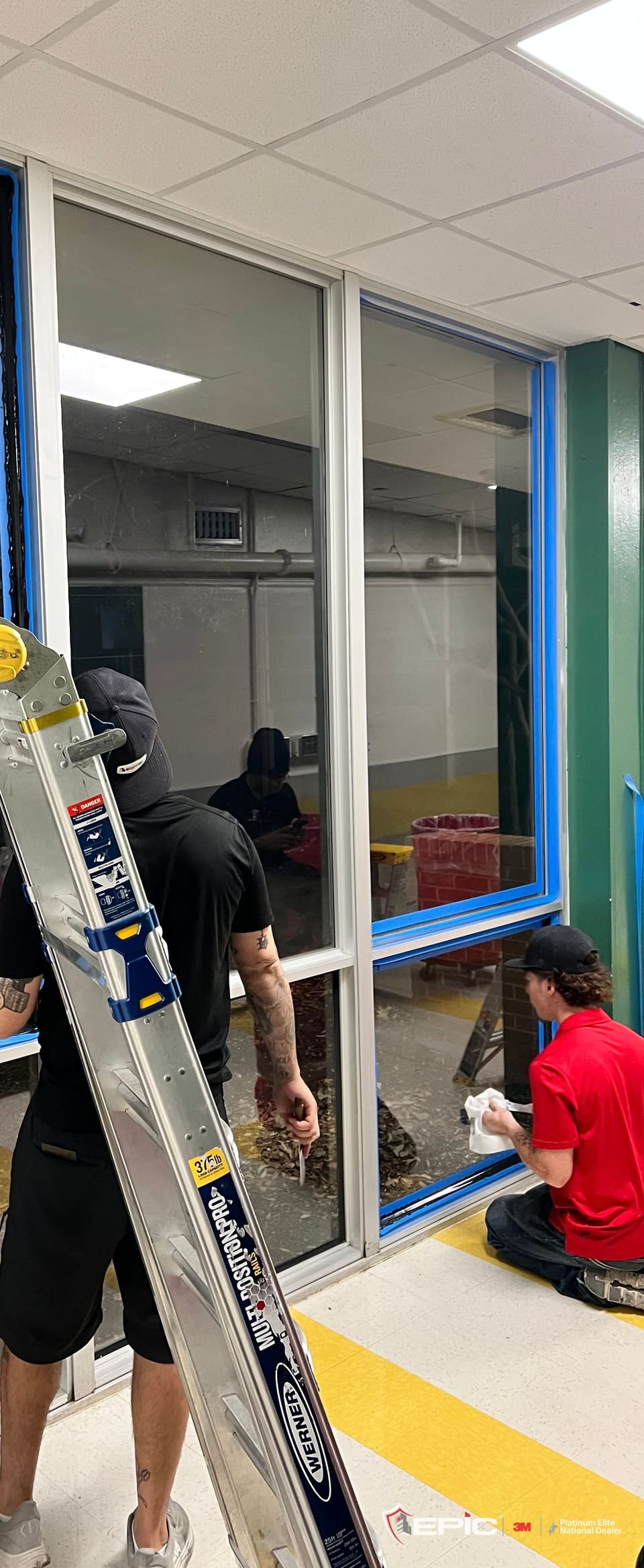 Cafeteria window lites at a Dallas ISD school taped in blue at night, the black 3M Impact Protection Attachment bead already down one frame edge, a Certified Installation Specialist kneeling at the sill