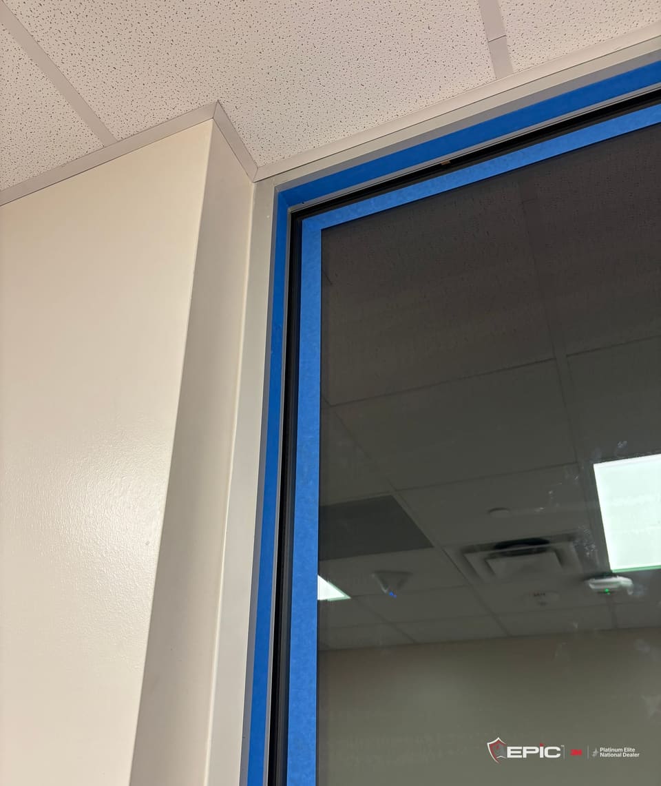 The top corner of an office window from the hall, the frame taped in blue, the film on