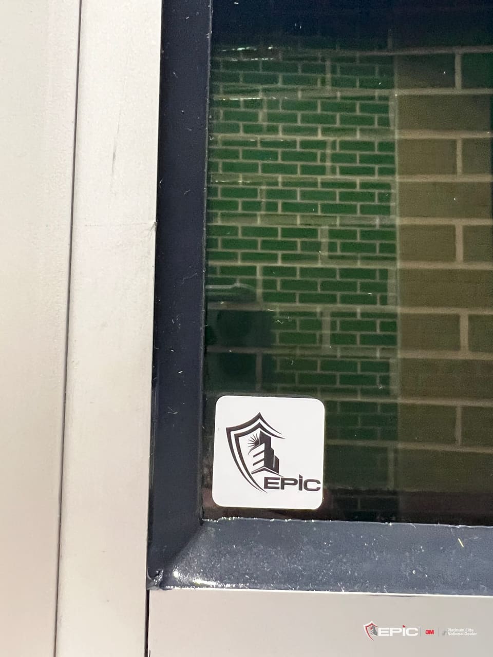 The lower corner of a window frame with the black 3M IPA bead run into the frame and a small Epic decal on the glass, green glazed brick beyond