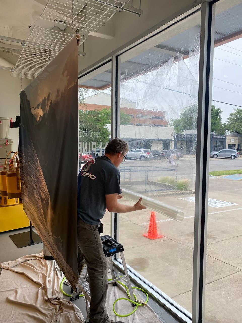 Epic Certified Installation Specialist applying security film to a tall storefront pane, the film roll and tools at his feet